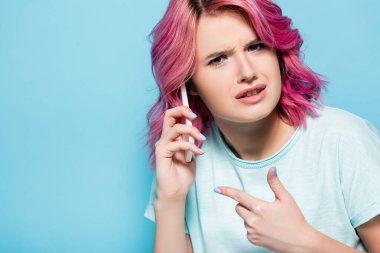 Kafası karışmış, pembe saçlı genç bir kadın mavi üzerinde izole akıllı telefonu işaret ediyor.