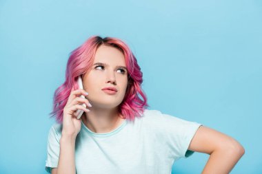 Pembe saçlı düşünceli genç kadın mavi arka planda akıllı telefondan konuşuyor.