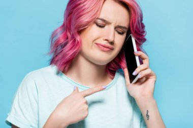 Kafası karışmış, pembe saçlı genç bir kadın mavi üzerinde izole akıllı telefonu işaret ediyor.