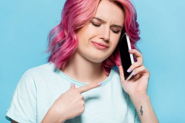 confused young woman with pink hair pointing at smartphone isolated on blue clipart