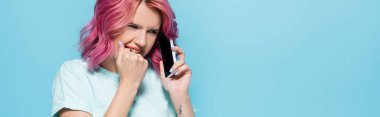 Pembe saçlı korkmuş genç kadın mavi arka planda akıllı telefondan konuşuyor, panoramik çekim.