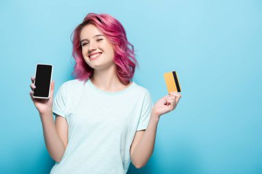 Pembe saçlı genç bir kadın elinde kredi kartı ve mavi arka planda akıllı telefon tutuyor.