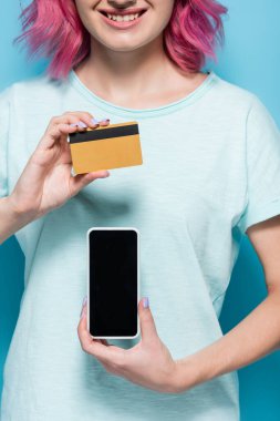 Pembe saçlı, elinde kredi kartı ve mavi arka planda akıllı telefonu olan genç bir kadının kısmi görüntüsü