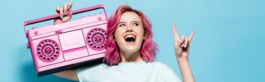 excited young woman with pink hair holding vintage tape recorder and showing rock sign on blue background, panoramic shot clipart