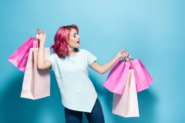 Pembe saçlı genç bir kadın mavi arka planda alışveriş torbaları tutuyor.