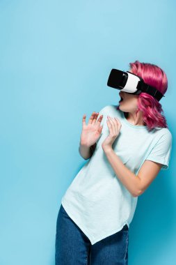 scared young woman with pink hair in vr headset on blue background