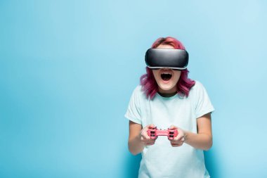 KYIV, UKRAINE - 29 Temmuz 2020: Mavi arka planda elinde joystick tutan pembe kulaklıklı, heyecanlı genç kadın