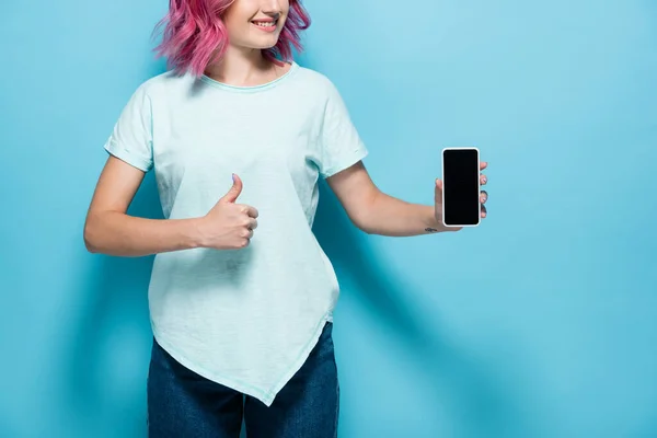 ピンク色の髪をした若い女性がスマートフォンを空の画面で持ち親指を青い背景に見せ — ストック写真