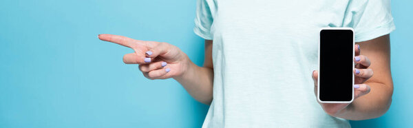 cropped view of young woman holding smartphone with blank screen and pointing aside on blue background, panoramic shot