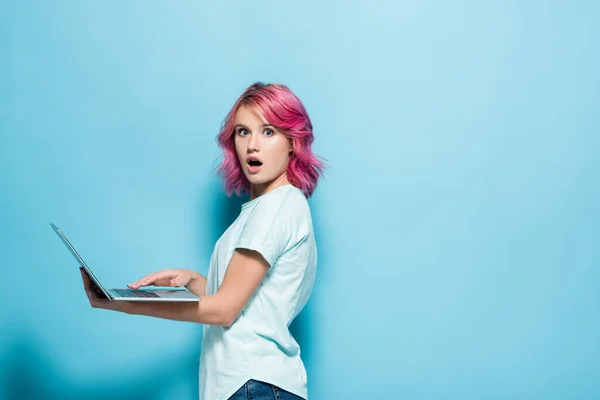 穿着粉色头发 手持蓝色背景笔记本电脑的年轻女子震惊的侧视图 — 图库照片