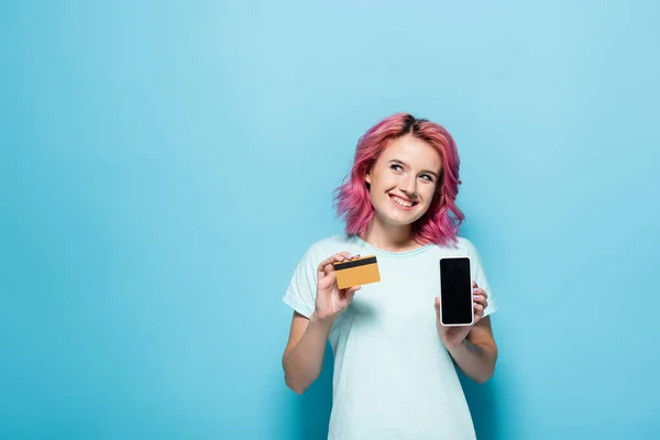 青い背景にピンクの髪をした若い女性がクレジットカードとスマートフォンを持っていて — ストック写真
