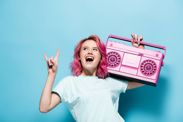 excited young woman with pink hair holding vintage tape recorder and showing rock sign on blue background