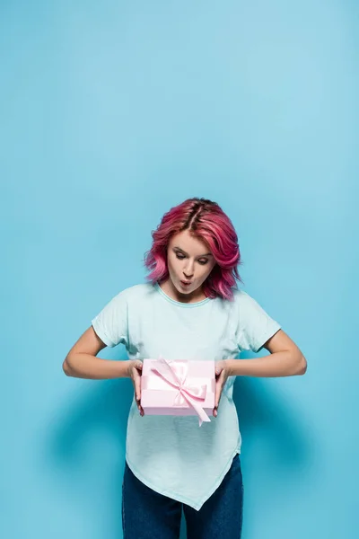 Zaskoczona Młoda Kobieta Różowymi Włosami Trzymając Pudełko Kokardą Niebieskim Tle — Zdjęcie stockowe