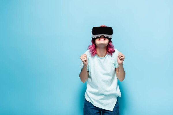 excited young woman with pink hair in vr headset showing yeah gesture on blue background