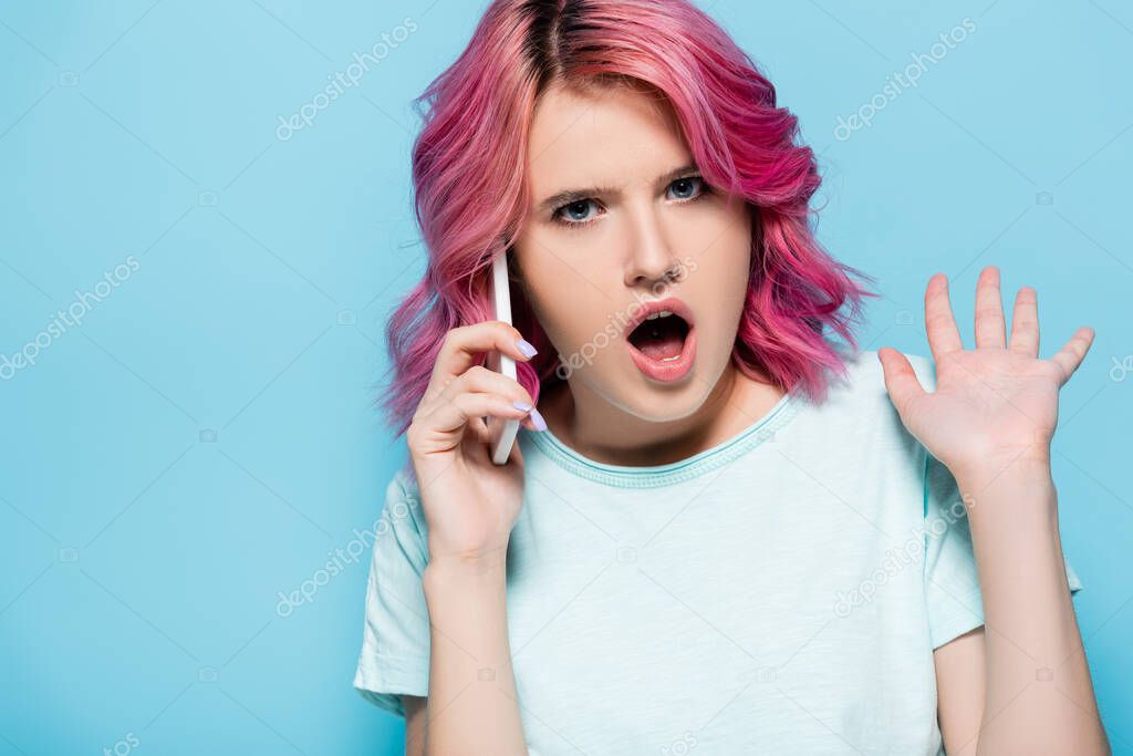 shocked young woman with pink hair talking on smartphone isolated on blue