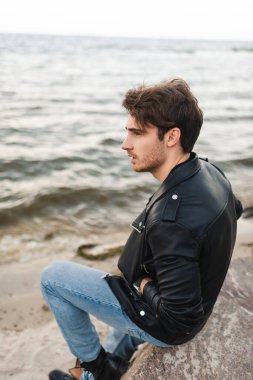 Deri ceketli ve kotlu bir adam sahilde taşın üzerinde oturuyor. 