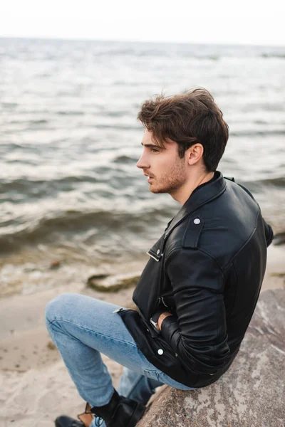 Man in leather jacket and jeans sitting on stone on beach