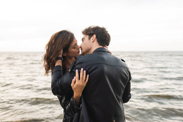 Мужчина в кожаной куртке целует девушку с морем на заднем плане 