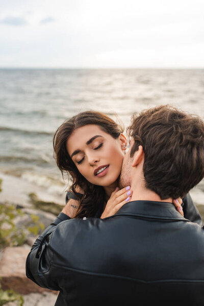 Мужчина в черной кожаной куртке целует подружку в шею возле моря на пляже 
