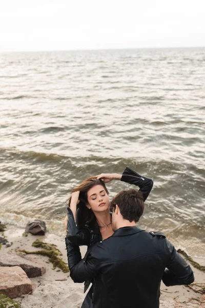 近岸海滩上一个穿着皮夹克亲吻女朋友的年轻人的高角照片 — 图库照片
