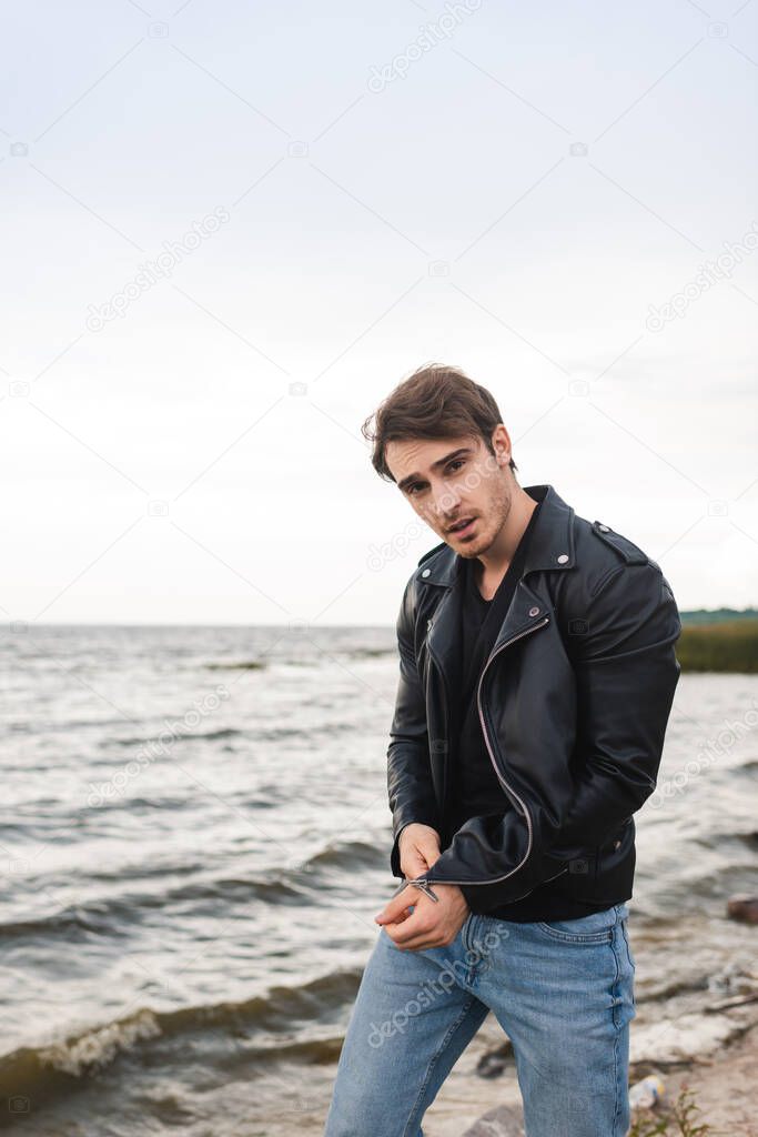 Young man in jeans and leather jacket looking at camera on beach 