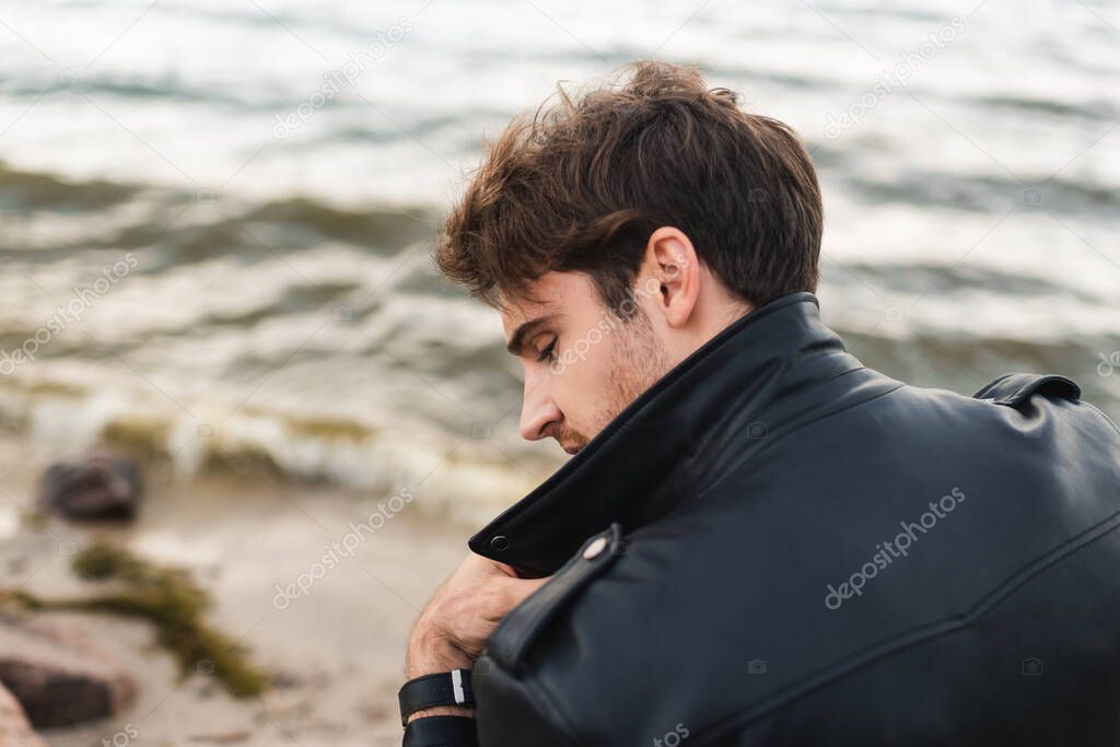 Back view of young man in black leather jacket on sea coast 