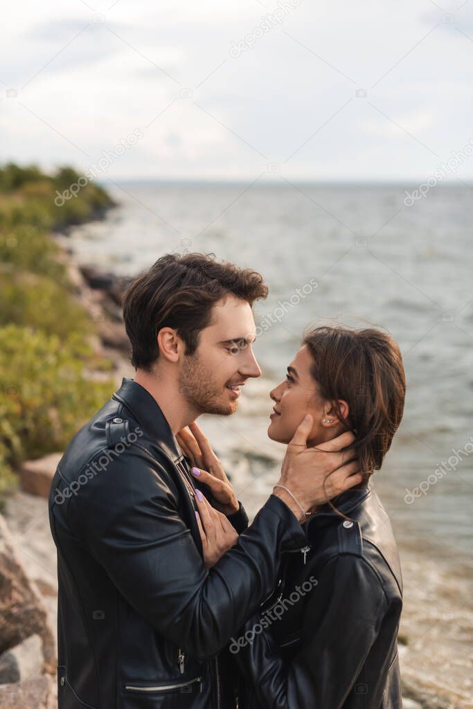 Side view of young couple in leather jackets touching and looking at each other near sea 