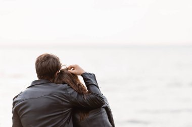 Deri ceketli bir adamın deniz kenarındaki kız arkadaşının saçlarına dokunuşunun arka görüntüsü. 