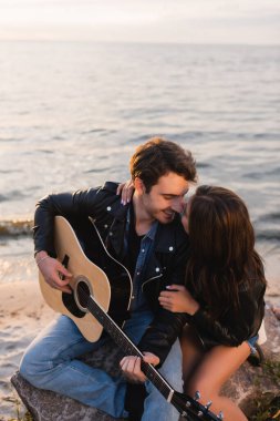Akustik gitarlı deri ceketli genç çift gün batımında sahilde oturuyor. 