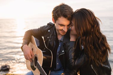 Akustik gitar çalan erkek arkadaşı kucaklayan bir kadın. Sahilde akşam vakti. 