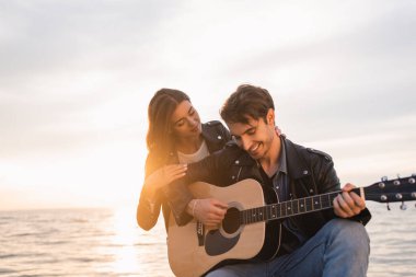 Sarılan erkek arkadaş, arka planda deniz kenarında akustik gitar çalıyor.