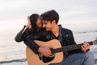 Gün batımında erkek arkadaşını öpen genç bir kadın deniz kenarında akustik gitar çalıyor. 