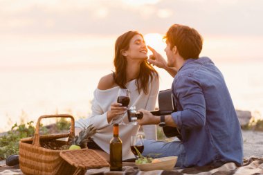 Akustik gitarlı bir adam gün batımında plajda bir bardak şarapla kız arkadaşının burnuna dokunuyor. 