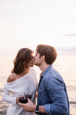 Selective focus of young man kissing nose of girlfriend with glass of wine near sea  clipart