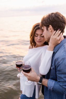 Elinde kırmızı şarap bardağı tutan esmer kadın ve deniz kenarında erkek arkadaşının boynu. 