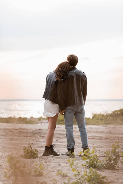 Back view of couple in leather jackets standing on sea beach at sunset 