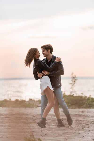 Selective focus of man in leather jacket embracing girlfriend on beach at sunset 