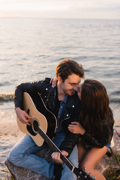Young couple in leather jackets with acoustic guitar sitting on beach during sunset 
