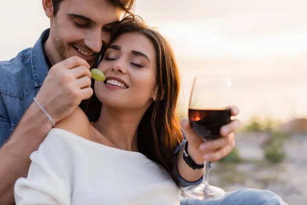 選択的焦点の男給餌ガールフレンドとともにブドウでワインのガラスを保持しながらビーチ — ストック写真