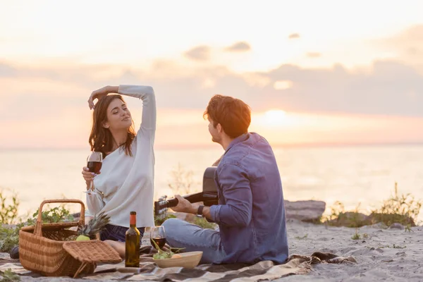 選択的焦点の男遊びますアコースティックギター近くガールフレンドとともにワインでビーチで夜 — ストック写真