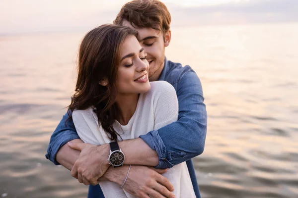 Jonge Man Knuffelen Brunette Vriendin Met Gesloten Ogen Naast Zee — Stockfoto