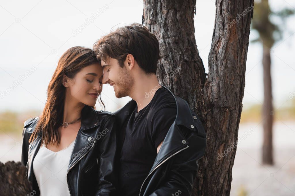 Selective focus of brunette woman in leather jacket standing near boyfriend with closed eyes in forest 