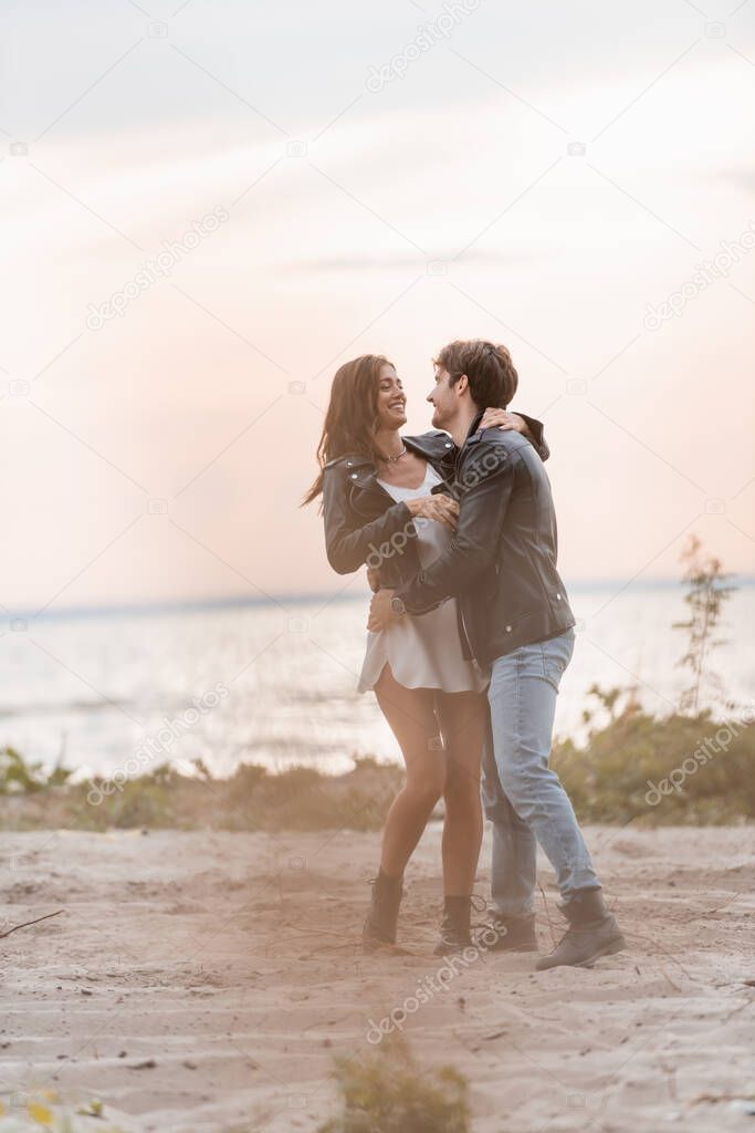 Selective focus of young couple in leather jackets hugging on beach at dawn 