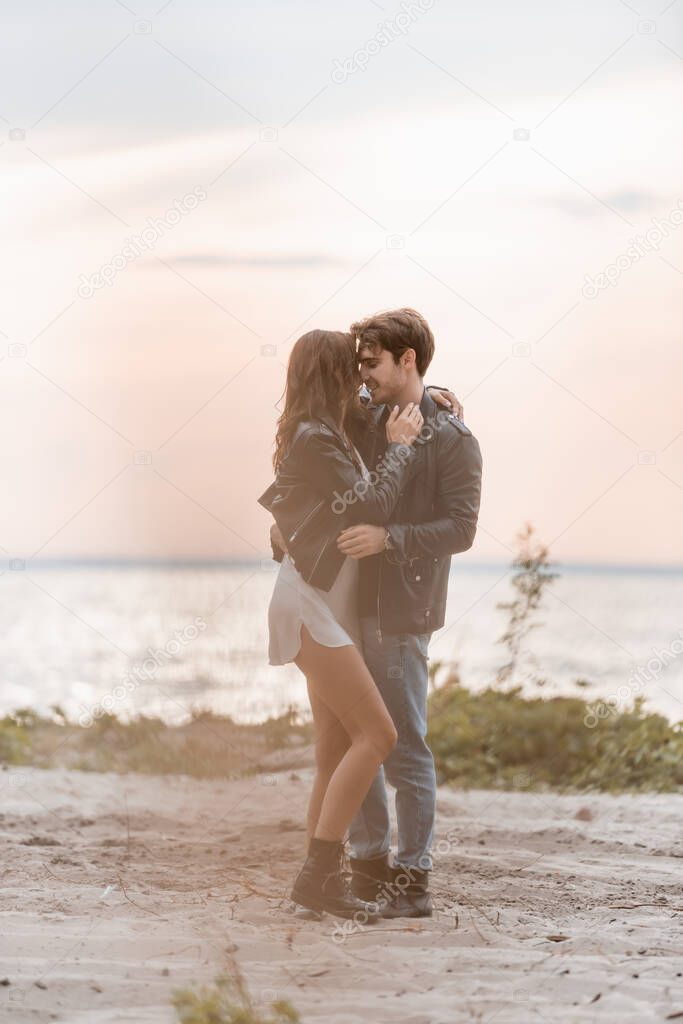 Selective focus of young man embracing girlfriend in dress and leather jacket on beach at sunset 