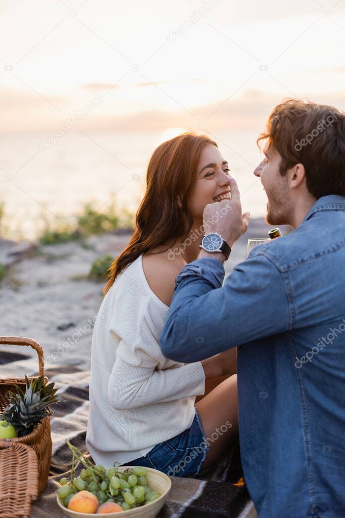 Selective focus of man with bottle of wine laughing while touching nose of girlfriend during picnic on beach 