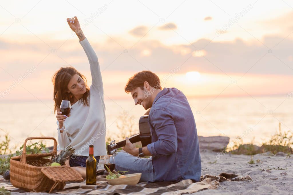 Selective focus of woman holding glass of wine near boyfriend playing acoustic guitar during picnic on beach 