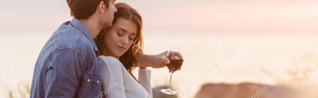 Website header of man hugging woman while holding glass of wine on beach at sunset 