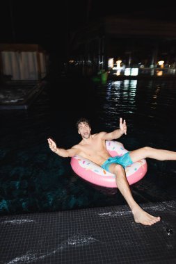 Selective focus of excited muscular man looking at camera on swim ring in pool at night  clipart