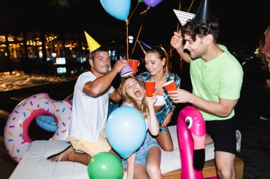 Friends in party caps holding disposable cups near swimming pool at night  clipart