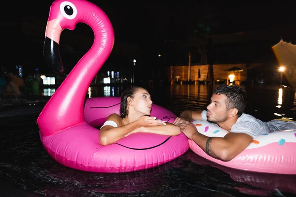 Young woman blowing air kiss to boyfriend while swimming in ring in pool at night